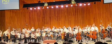 The Portland Symphony Orchestra on stage at the OOB Seaside Pavilion. The musicians all wear white shirts and dark pants. a screen in the upper left of the image shows the PSO logo.