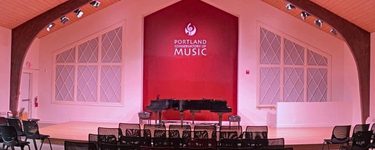 The interior of the portland conservatory of music. the low stage is behind some black chairs, and has a pair of black grand pianos on it. There is a red panel with the PCM logo on it behind the stage, and false windows to either side. PCM is a converted church, so it has something of a cathedral feel to it.
