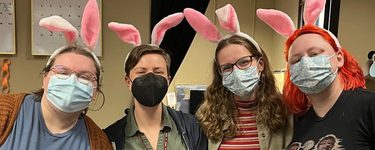 four white women wearing face masks and bunny ears. Behind them above a doorway is a sign with reads 