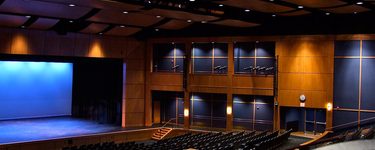 The interior of the Westbrook performing arts center from the back of the house. in the bottom/foreground of the image you can see the plastic backs of two chairs. the stage is on the left side of the stage, with a cyc lit up blue as a backdrop. the main focus o the image is the right wall of the theatre which has nice wood paneling with dark blue panels and lighting instruments hung up.